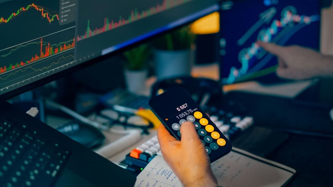 Candlestick charts on two monitors while a trader works a figure out on a phone calculator